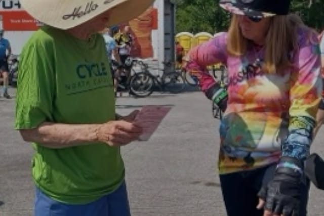 At a cycling event, a rider in a tie-dye jersey and wrist guards talks with a volunteer reading a flyer.