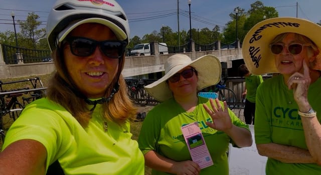 A rider in a white helmet takes a selfie with two Cycle North Carolina volunteers, one holding a printed flyer.